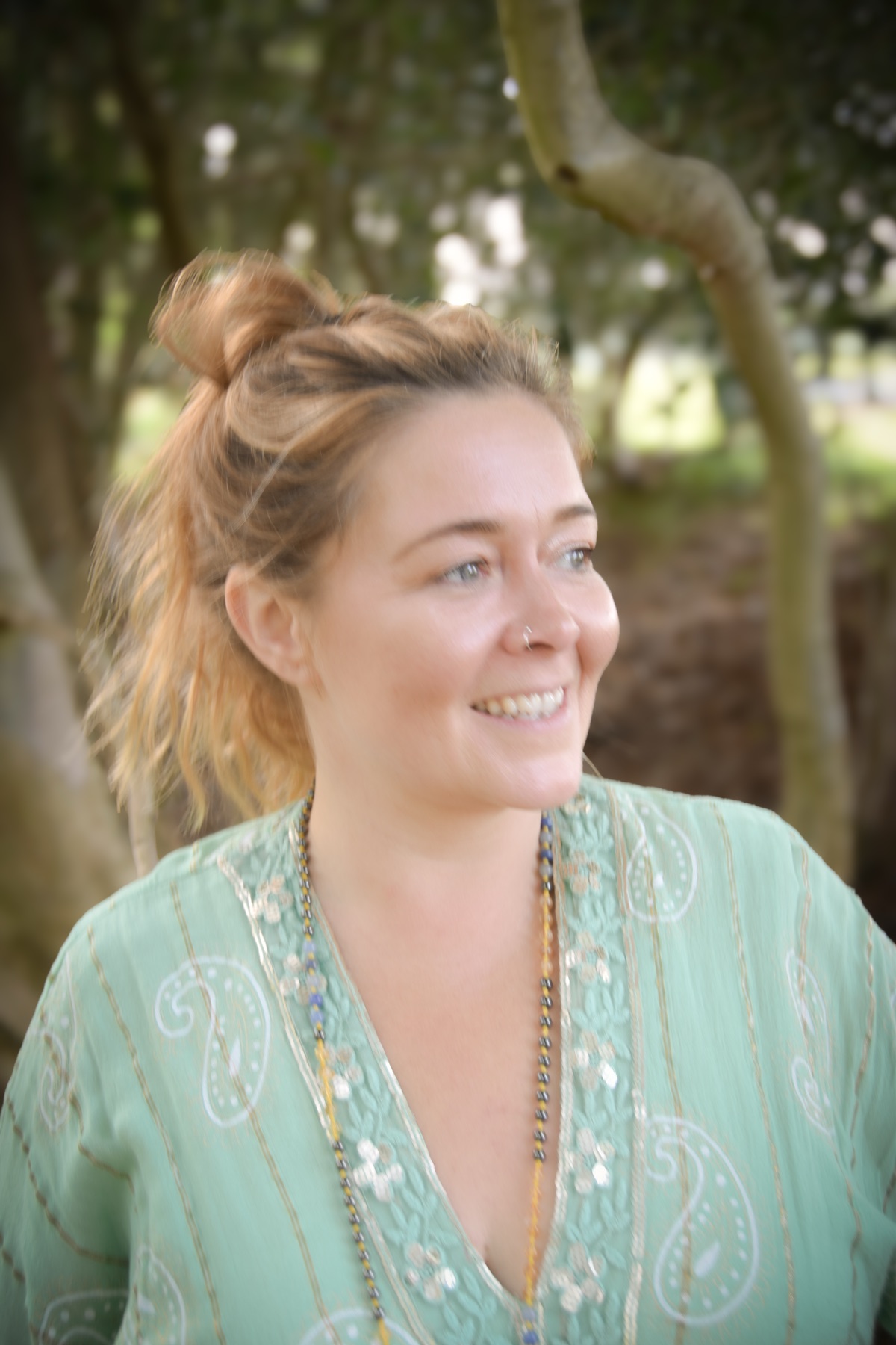 Jade in a sea-green paisley top, smiling and looking to one side beneath a tree