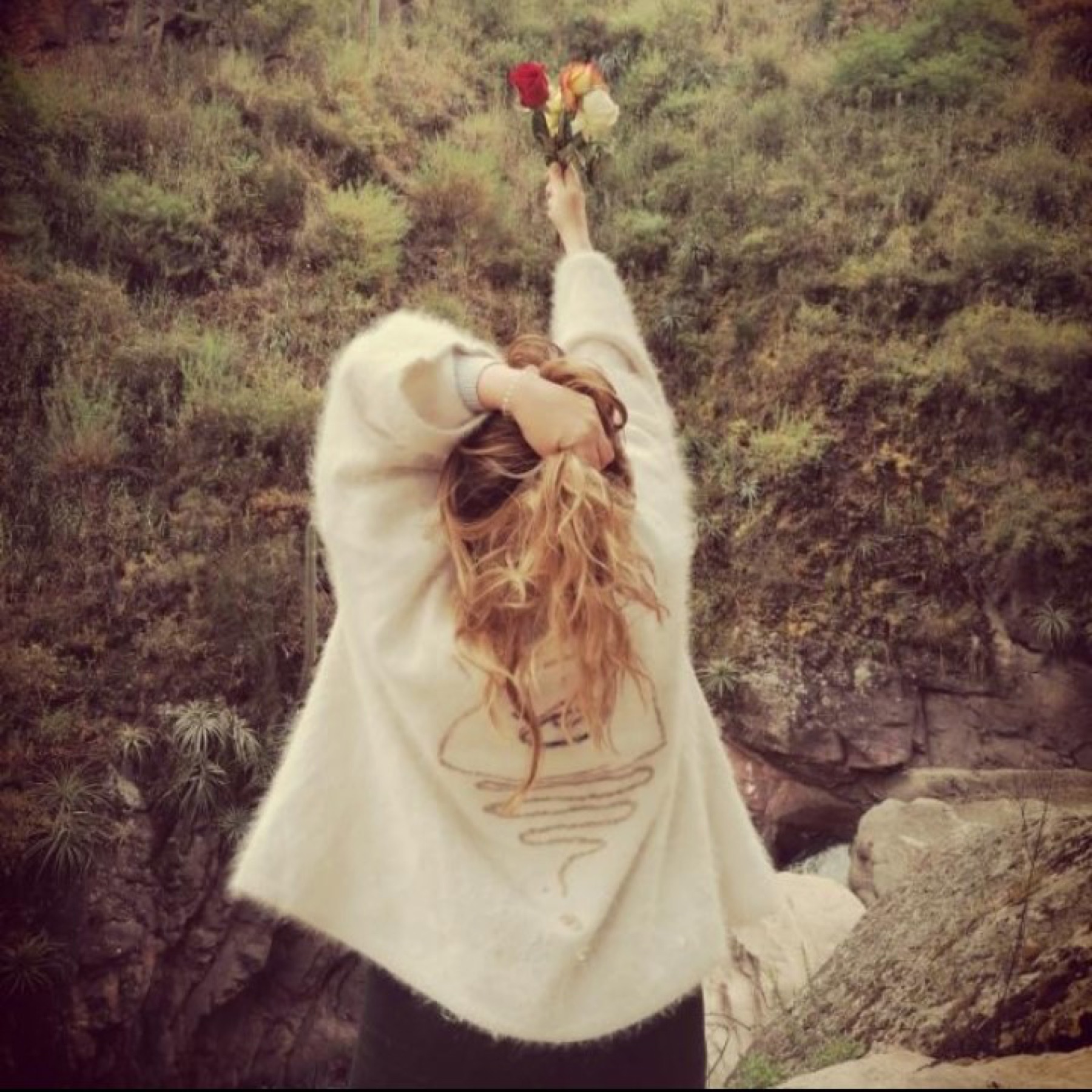 A woman in a cream jumper raising a bunch of flowers to the sky beside a stream